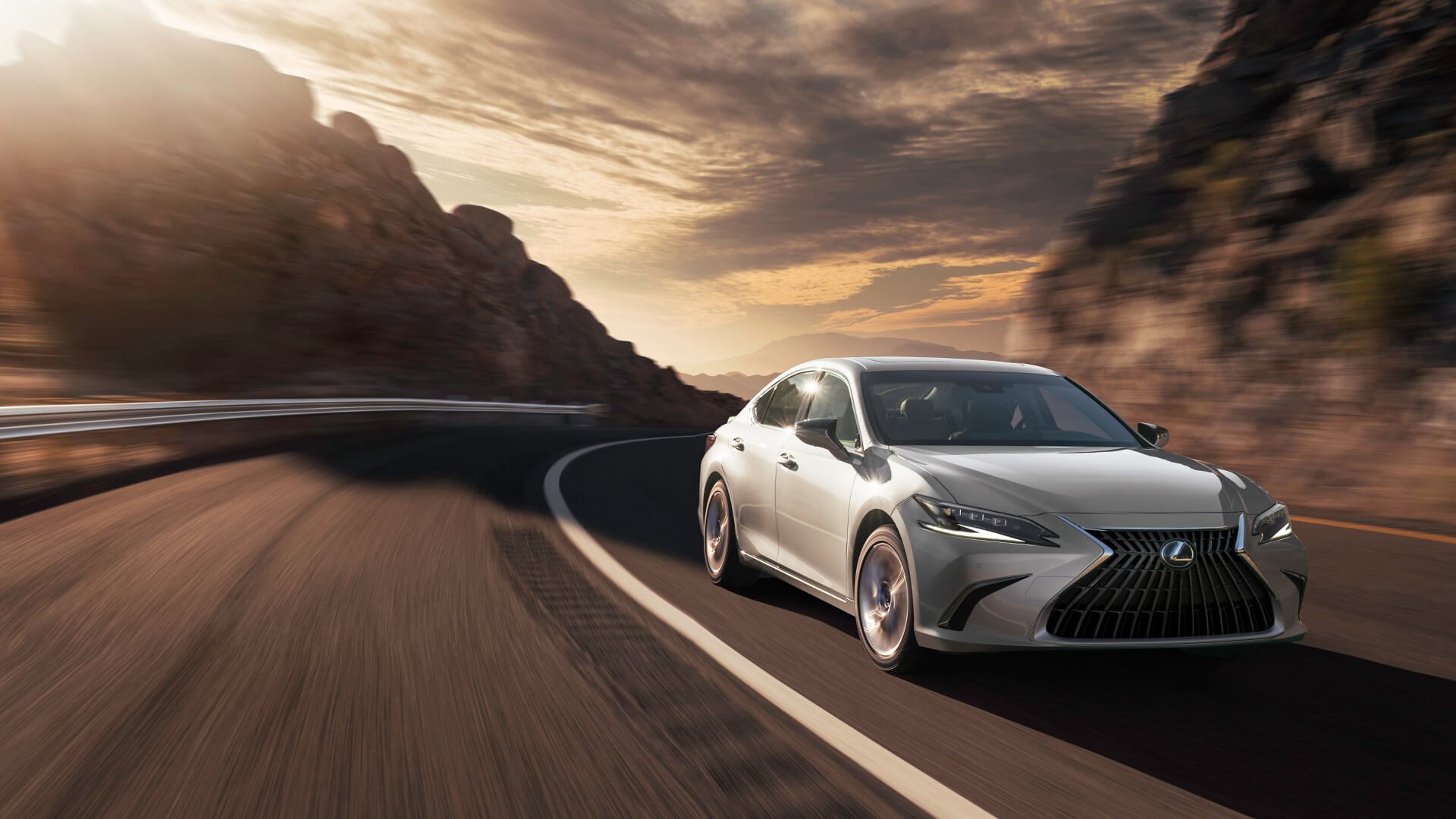 Silver Lexus ES is being driven on the road with a mountainous backdrop.